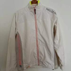 Vit vindjacka från Helly Hansen - Stilren vit vindjacka från Helly Hansen med hel dragkedja framtill och diskreta röda detaljer. Jackan har trycket 'SPEED PERFORMANCE' på ena ärmen och logga på axeln. Lätt material och hög krage med gul detalj på insidan. Längd: 70cm, P2P: 50cm