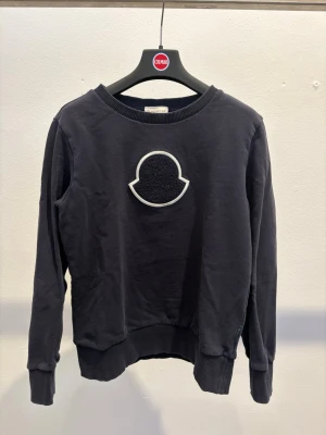 Svart hoodie från Moncler - Svart hoodie från Moncler med stort broderat logomärke på bröstet. Klassisk rund halsringning och ribbade muddar vid ärmslut och nederkant. Perfekt för en stilren och avslappnad look. Något urtvättad.