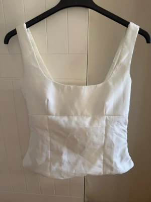 Vit linnetopp med rosett i ryggen - Stilren vit linnetopp med breda axelband och fyrkantig ringning. Toppen har en snygg rosettdetalj i ryggen som ger en extra touch. Perfekt att matcha med jeans eller kjol för en fräsch look. Materialet känns svalt och lätt. Köpt på plick innan och har inget märke eller lapp för storlek i sig. Jag skulle säga att de är en S/M