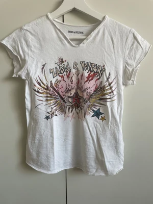 Vit t-shirt från Zadig & Voltaire - Säljer en vit t-shirt från Zadig & Voltaire med coolt grafiskt tryck framtill i flera färger och stjärndetaljer. T-shirten har korta ärmar, rund halsringning med rå kant och två dekorativa knappar vid halsen. Perfekt för en edgy och avslappnad stil.