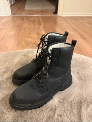 Svarta fodrade kängor med snörning - Svarta kängor i skinnimitation från DinSko med snörning och grov sula. Insidan är fodrad med vitt, mjukt foder som håller värmen. Perfekta för kalla dagar och har en klassisk, stilren look.