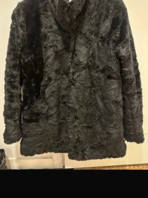 Svart fuskpälsjacka med krage stl S - Mysig svart fuskpälsjacka med hög krage och rak passform. Jackan har en fluffig och mjuk yta som ger en lyxig känsla. Perfekt för kalla dagar när du vill hålla dig varm och snygg. Stängning framtill och helsvart design.