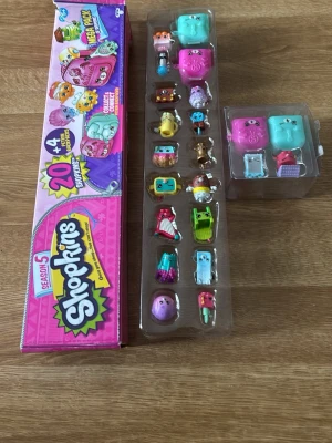 Shopkins Säsong 5 samlarfigurer - Shopkins Säsong 5 samlarfigurer i originalförpackning. Paketet innehåller 20 små färgglada plastfigurer i olika former och färger, samt tillbehör och två rosa och turkosa förvaringsboxar. Perfekt för dig som gillar att samla och leka med Shopkins.