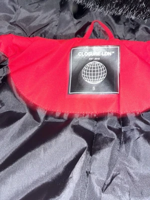 Röd vindjacka från Closure LDN - Snygg röd vindjacka från Closure LDN med svart foder och huva med fuskpäls. Jackan har två bröstfickor med dragkedja och två stora fickor med lock. Svarta muddar vid ärmslut och midja ger en schysst passform.