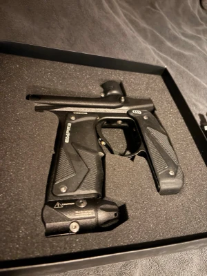 Empire mini Gs paintballmarkör - Säljer en svart paintballmarkör från Empire med greppvänliga handtag och robust design i metall och plast. Markören har detaljerade mönster på handtaget och en tryckmätare framtill. Perfekt för dig som gillar paintball och vill ha bra utrustning.