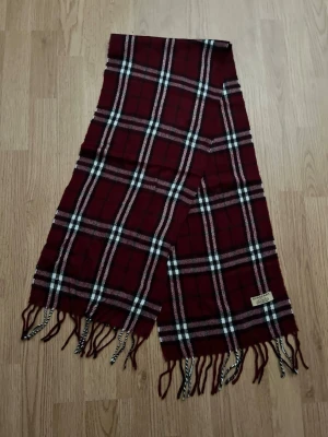Rutig vinröd halsduk från Burberry - Snygg vinröd halsduk från Burberry med klassiskt rutmönster i svart, vitt och beige. Halsduken har fransar i ändarna och är tillverkad i mjukt material som känns lyxigt och stilrent. Perfekt accessoar för att lyfta din outfit. Kostar ca 5000kr nytt. Pris går att diskuteras vid snabb deal