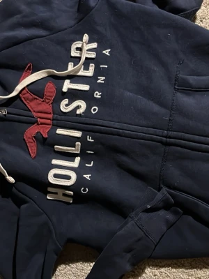 Mörkblå Hollister hoodie med dragkedja - Mörkblå hoodie från Hollister med vit dragkedja och snören i huvan. Stor röd logga och broderad text på framsidan. Mjuk insida i fleece och klassisk känguruficka framtill. Perfekt för en avslappnad och cool stil. Bra skick och jag postar samma dag. Laidback/ Avicii stil🔥