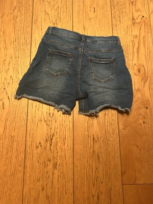 Blå jeansshorts med fransig kant - Säljer ett par klassiska blå jeansshorts med fransig, rå kant. Shortsen har hög midja och är tillverkade i ett slitstarkt denimtyg. Perfekta för varma dagar och enkel att matcha med olika stilar.