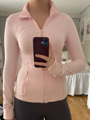 Lululemon define jacket - Snygg ljusrosa träningsjacka med hel dragkedja framtill och hög krage. Figurnära passform och långa ärmar med tumhål. Två fickor med dragkedja på sidorna. Perfekt för gymmet eller löprundan.
