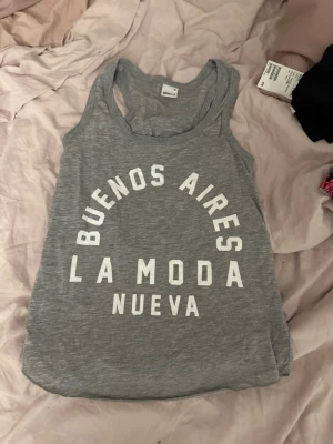 Grå linnetopp med tryck - Grå ärmlös linnetopp med vit text 'Buenos Aires La Moda Nueva' framtill. Avslappnad passform och rund halsringning, perfekt för en chill look.