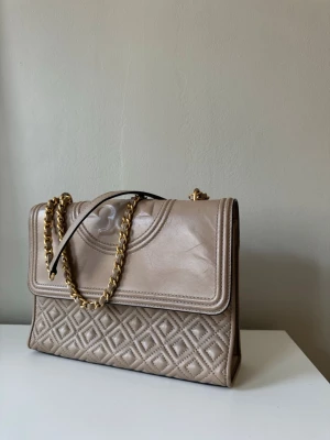 Tory Burch väska – beige 🤍 - Säljer en äkta Tory Burch-väska i beige, använd sparsamt under några år. Väskan är i fint skick men har en mindre repa (se bild), därav priset. I övrigt mycket väl omhändertagen.  En klassisk och tidlös modell som passar perfekt till både vardag och fest. Lättmatchad färg 🌟  ✔️ Äkta ✔️ Sparsamt använd ✔️ Mindre repa finns ✔️ Rök- & djurfritt hem  Skickas snabbt 💌 Hör av dig vid frågor eller om du vill ha fler bilder!