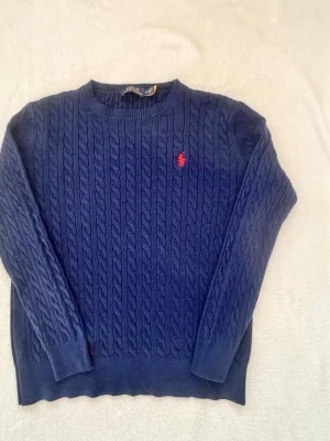 Mörkblå kabelstickad tröja Polo Ralph Lauren - Kabelstickad mörkblå tröja från Polo Ralph Lauren med rund halsringning och röd broderad logga på bröstet. Tröjan har ribbade muddar vid ärmslut och nederkant. Klassisk och tidlös design i mjukt material. 