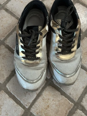 Michael Kors sneakers i guld och vitt - Snygga sneakers från Michael Kors i vitt och guld med svarta detaljer. Ovandelen har mesh och metallicpaneler, samt svarta snören med vita inslag. Perfekta för dig som vill sticka ut med en lyxig touch. Välanvända men fortfarande mer att ge. Nypris 2300:-
