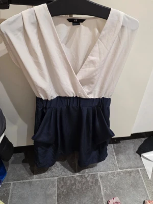 Vit och marinblå omlottklänning H&M - Snygg kortklänning från H&M i omlottmodell med vit överdel och marinblå kjol. Klänningen har markerad midja och volangdetaljer på kjolen. Perfekt för dig som gillar stilrena kontraster och en feminin siluett.