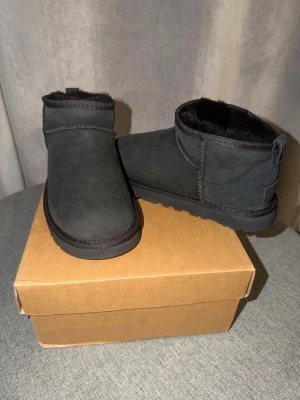 Svarta UGG tofflor i mocka - Säljer ett par svarta UGG tofflor i mjuk mocka med fluffigt foder och robust gummisula. Klassisk låg modell som är både stilren och bekväm. Perfekta för att hålla fötterna varma under kalla dagar.