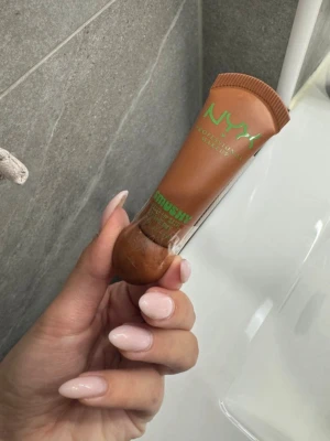 NYX Mushy läppbalsam brun tub - NYX Professional Makeup Mushy läppbalsam i en mjuk tub med brun färg och grön text. Tuben är enkel att använda och perfekt att ha med sig. Ger återfuktning till läpparna med en krämig formula och naturlig finish.