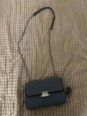 Mörkblå axelväska med kedjerem - Snygg mörkblå axelväska i slät konstläder med silverfärgad kedjerem och metallspänne framtill. Väskan har en tofsdetalj på sidan och stilren, rektangulär form. Perfekt för dig som gillar minimalistisk design och vill ha en väska som sticker ut.