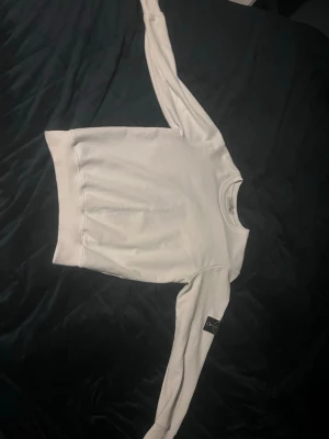 Vit sweatshirt från Stone Island - Säljer en clean vit sweatshirt från Stone Island med klassisk rund hals och ribbade muddar. Tröjan har den ikoniska Stone Island-loggan på ärmen och är gjord i mjukt bomullsmaterial. Perfekt för dig som gillar stilrena och exklusiva plagg.