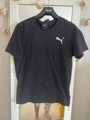 Svart Puma t-shirt med logga - En svart t-shirt från Puma med klassisk vit logga på bröstet. T-shirten har rund halsringning och korta ärmar. Perfekt för en sportig och avslappnad stil. Tillverkad i mjuk bomull för skön komfort.