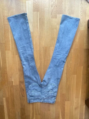 Gråa bootcut jeans med detaljer - Säljer ett par gråa bootcut jeans med unika bakfickor som har dekorativa sömmar och lock. Jeansen har en avslappnad passform och är tillverkade i mjukt denimtyg. Perfekta för dig som gillar en retro och avslappnad stil.En av knapparna saknas på en av fickorna