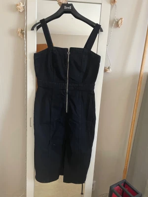 Svart jeansklännig  med dragkedja - Snygg svart byxdress med breda axelband och markerad midja. Dragkedja framtill som detalj och raka ben. Perfekt för dig som gillar en stilren look med edge. Materialet ser ut att vara kraftigt bomullstyg eller denim.klänning är som jeans .