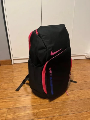 Nike Elite ryggsäck - Snygg svart ryggsäck från Nike med rosa dragkedjor och logga. Framsidan har en cool grafisk detalj i rosa och blått. Ryggsäcken har flera fack och justerbara axelremmar. Perfekt för skolan eller träningen.