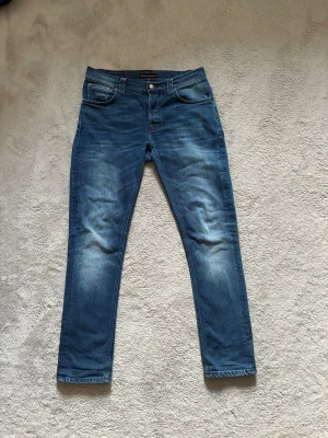 Blå slim fit jeans från Nudie Jeans - Säljer ett par blå slim fit jeans från Nudie Jeans med klassisk femficksmodell och snygga slitningar framtill. Jeansen har normal midja, raka ben och är tillverkade i mjuk denim. Bakfickorna har Nudie Jeans karakteristiska broderi.