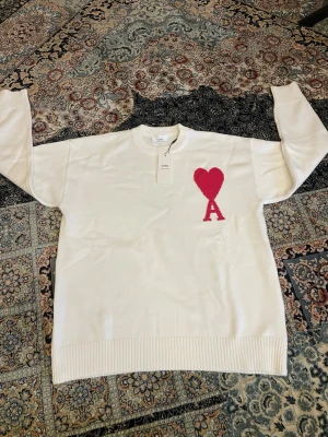 Vit tröja  Ami Paris - Vit stickad tröja från Ami Paris med rund halsringning och ribbade muddar. På bröstet finns ett rött hjärta med bokstaven 'A' under, vilket är Ami Paris ikoniska logga. Tröjan har en clean look och är perfekt för dig som gillar stilrena plagg.