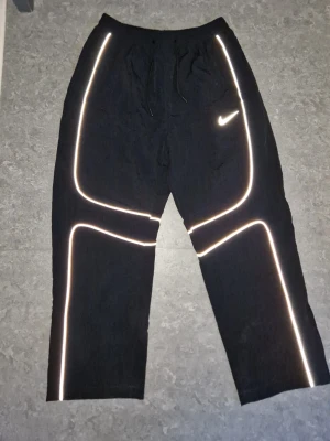 Nike X Corteiz NRG Byxor - Säljer ett par svarta träningsbyxor från Nike och Corteiz med reflexdetaljer längs benen och runt fickorna. Byxorna har resår i midjan och dragsko för bästa passform. Materialet är mjukt och bekvämt, perfekt för träning eller chill. 