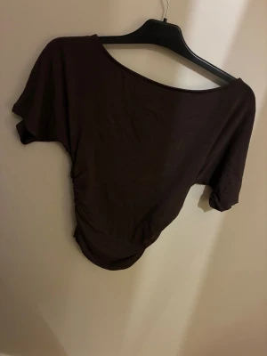 Brun asymmetrisk topp med rynk - Snygg brun topp med asymmetrisk halsringning och kort ärm. Toppen har rynkade detaljer på ena sidan vilket ger en cool och trendig look. Perfekt att styla med jeans eller kjol för en avslappnad vibe.