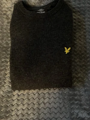 Svart stickad tröja från Lyle & Scott - Mörkgrå stickad tröja från Lyle & Scott med rund halsringning och deras klassiska gula fågel-logga broderad på bröstet. Tröjan har långa ärmar och ribbade muddar vid ärmslut och nederkant. Perfekt för lager-på-lager-stil.