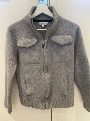 Stickad kofta från Giovani 🇮🇹 - Snygg grå kofta från Giovani 🇮🇹 med hög krage och två bröstfickor. Krympte i tvätten därav PRISET. Tröjan är stickad med ribbade muddar vid ärmslut och nederkant. Perfekt för lager-på-lager och enkel att matcha med andra plagg. 100% Ull