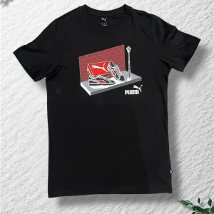 Svart Puma t-shirt med tryck - Svart t-shirt från Puma med coolt grafiskt tryck framtill som visar sneakers, tegelvägg och gatlykta. Klassisk passform och rund halsringning. Perfekt för dig som gillar streetstyle och sportiga märken.