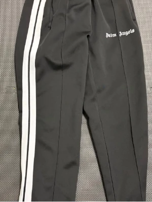 Palm Angels svarta mjukisbyxor - Svarta mjukisbyxor från Palm Angels med vita dubbla sidoremsor och logga tryckt på vänster ben. Byxorna har elastisk midja och är tillverkade i ett mjukt, sportigt material som känns skönt mot huden. Perfekta för en avslappnad och trendig streetwear-look.