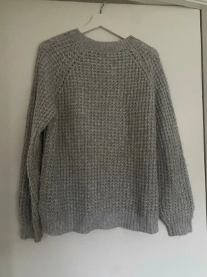 Ljusgrå stickad tröja - Mysig ljusgrå stickad tröja med grov struktur och rund hals. Tröjan har långa ärmar och ribbade muddar vid ärmslut och nederkant. Perfekt för kyliga dagar och enkel att matcha med olika outfits.