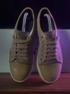 Beige sneakers från Lanvin i mocka - Säljer ett par stilrena beige sneakers från Lanvin med ovandel i mocka och tåparti i skinn. Skorna har klassisk snörning och platt sula, perfekt för dig som gillar en clean och lyxig look. Diskret Lanvin-logga under sulan och snygga detaljer i ton-i-ton.
