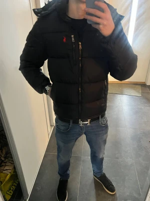 Ralph Lauren jacka  - Säljer en svart pufferjacka från Polo Ralph Lauren med röd logga på bröstet. Jackan har dragkedja framtill, flera fickor och en huva. Perfekt för kalla dagar och riktigt snygg till jeans eller streetwear. Materialet är slitstarkt och håller värmen bra.