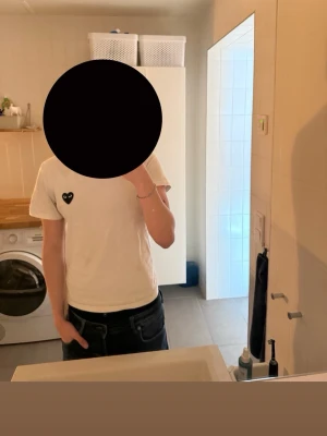 Vit t-shirt från Comme des Garçons - Säljer en klassisk vit t-shirt från Comme des Garçons med det ikoniska svarta hjärtat på bröstet. T-shirten har korta ärmar och en normal passform. Perfekt till jeans eller shorts för en clean och stilren look.