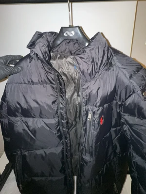 Svart dunjacka från Polo Ralph Lauren - Snygg svart dunjacka från Polo Ralph Lauren med klassisk röd logga på bröstet. Jackan har huva, dragkedja framtill och är vadderad för extra värme. Perfekt för kalla vinterdagar och har en stilren look.