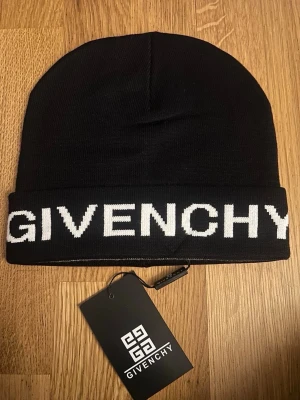 Svart-grå mössa från Givenchy - Snygg svart-grå mössa från Givenchy med vit logotyp broderad runt uppviket. Klassisk modell i mjukt stickat material som sitter skönt på huvudet. Perfekt accessoar för att lyfta din streetwear-look.