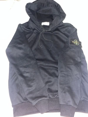 Svart hoodie från Stone Island - Säljer en svart hoodie från Stone Island med klassisk patch på vänster ärm. Hoodien har dragsko i huvan och ribbade muddar vid ärmslut och nederkant. Tillverkad i mjukt bomullsmaterial som är skönt att bära. Perfekt för dig som gillar streetwear och vill ha en stilren look.