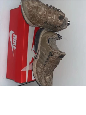 Nike  presto  - Nike sneakers med beige och brunt camomönster, lågt skaft och svart sula. Skorna har mesh- och syntetmaterial, rund tå och klassisk Nike-logga på hälen. Sitter på en röd Nike-låda. Perfekta för dig som gillar streetwear och unika detaljer.
