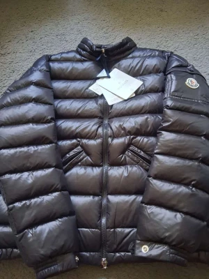 Svart dunjacka från Moncler - Snygg svart dunjacka från Moncler med glansig finish och klassisk quiltad design. Jackan har två snedställda fickor med dragkedja, hög krage och Moncler-logga på ärmen. Perfekt för kalla vinterdagar och riktigt trendig look. Storlek 2/M men passar som 1/S