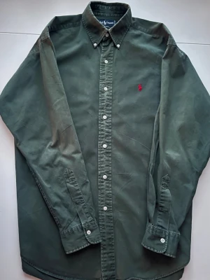 Grön skjorta från Ralph Lauren M - Klassisk grön skjorta från Ralph Lauren i modellen Blaire. Skjortan har knappar hela vägen, button down-krage och den ikoniska röda broderade loggan på bröstet. Tillverkad i 100% bomull och har långa ärmar. Perfekt för en stilren och avslappnad look.