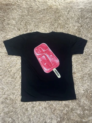 Svart Stüssy t-shirt med glassmotiv - Svart t-shirt från Stüssy med en stor rosa glass på ryggen och Stüssy-logga på bröstet. T-shirten har klassisk passform och är tillverkad i mjuk bomull. Perfekt för dig som gillar streetwear och coola tryck.