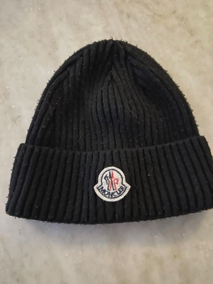 Svart Moncler ribbstickad mössa - Snygg svart ribbstickad mössa från Moncler med uppvikt kant och klassisk Moncler-logga framtill. Perfekt accessoar för kalla dagar och enkel att matcha med olika outfits. Materialet är mjukt och värmande.