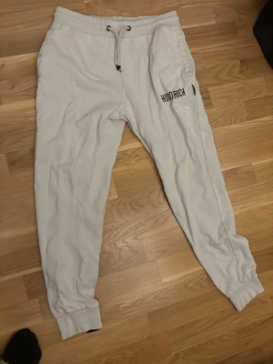 Grå mjukisbyxor från Hoodrich - Snygga grå mjukisbyxor från Hoodrich med svart logga på låret och dragkedjeficka. Byxorna har resår och dragsko i midjan samt muddar vid bensluten. Perfekta för en chill och avslappnad stil.