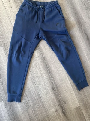 Blå Nike mjukisbyxor med dragsko - Snygga blå mjukisbyxor från Nike med resår och dragsko i midjan. Byxorna har ribbade muddar vid bensluten, sidofickor och en extra ficka med dragkedja bak. Perfekta för en avslappnad och sportig stil.