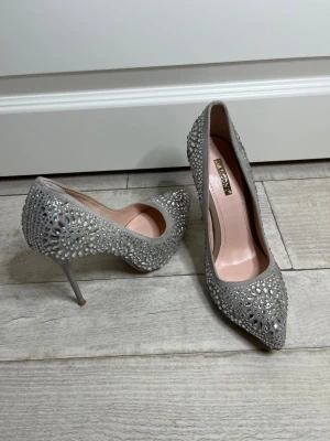 Grå pumps med strass från Basconi - Supersnygga grå pumps från Basconi i storlek 36 men passar också 37, skorna har en spetsig tå och höga klackar. Skorna är täckta av glittrande strass och har en elegant utskärning på sidan. Insidan är ljusrosa och materialet ser ut att vara mocka.