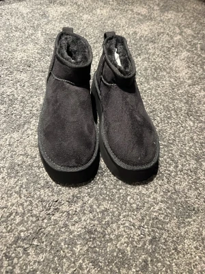 Svarta fluffiga slip-in tofflor - Säljer ett par svarta slip-in tofflor med högre skaft och mjukt, fluffigt foder. Yttermaterialet är i mockaimitation och sulan är tjock och robust. Perfekta för att hålla fötterna varma hemma eller på språng.